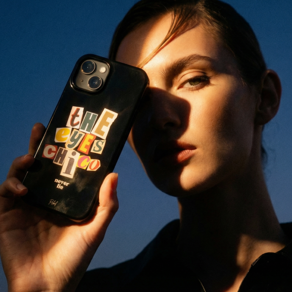 Person holding a phone case with 'The Eyes Choco' design against a blue sky.